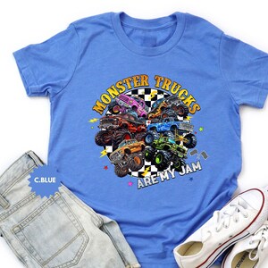 May include: A light blue t-shirt featuring a colorful graphic of monster trucks. The design includes the text "MONSTER TRUCKS ARE MY JAM" in yellow and orange. The shirt is paired with light wash denim jeans and white sneakers.