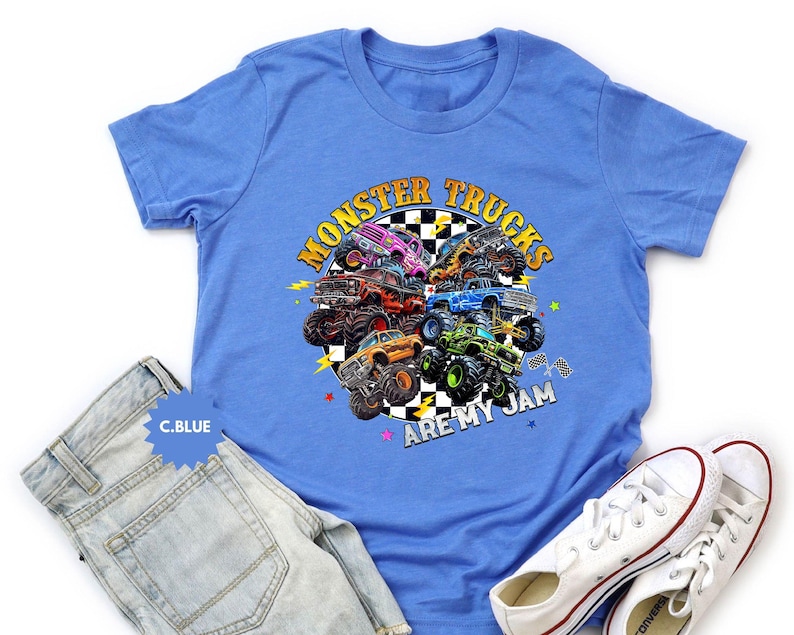 May include: A light blue t-shirt featuring a colorful graphic of monster trucks. The design includes the text "MONSTER TRUCKS ARE MY JAM" in yellow and orange. The shirt is paired with light wash denim jeans and white sneakers.