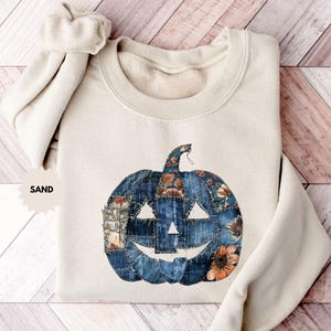 Op de afbeelding: Een zandkleurige sweatshirt met een groot, geappliqueerd pompoenontwerp. De pompoen is gemaakt van denim en bloemenstof patches, met een lachend gezicht uitgesneden. Het woord "SAND" staat links op de sweatshirt.