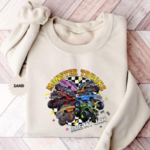 May include: Sand-colored sweatshirt featuring a colorful graphic of monster trucks. The design includes the text "MONSTER TRUCKS ARE MY JAM" in a circular arrangement. The sweatshirt has a crew neck and long sleeves.