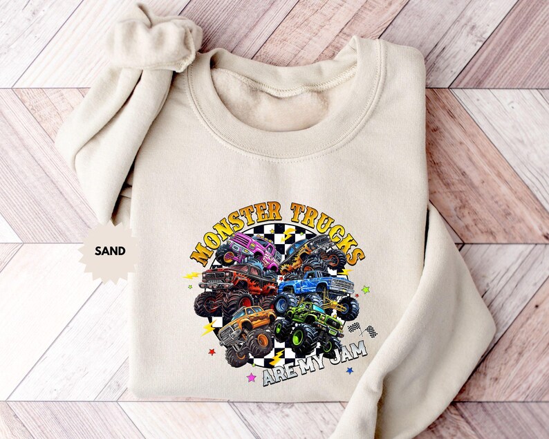 May include: Sand-colored sweatshirt featuring a colorful graphic of monster trucks. The design includes the text "MONSTER TRUCKS ARE MY JAM" in a circular arrangement. The sweatshirt has a crew neck and long sleeves.