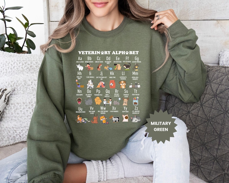 May include: Military green sweatshirt featuring the "Veterinary Alphabet" with various dog breeds and related items for each letter. The sweatshirt has long sleeves and a crew neck. The text "MILITARY GREEN" is printed on the bottom right.