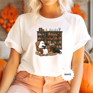 May include: White t-shirt with a graphic of a ghost reading a book in a library with a black cat. The text "WHITE" is visible in the bottom right corner of the image.