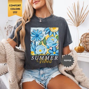 Peut inclure: Un t-shirt gris fonc&eacute; avec un motif de fleurs color&eacute;es et le texte "Summer Vibes" imprim&eacute; sur le devant. Le t-shirt a des manches courtes et un col rond.