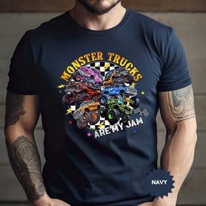May include: Navy blue t-shirt featuring a colorful graphic of monster trucks. The design includes the text "MONSTER TRUCKS ARE MY JAM" in a circular arrangement. The shirt is a solid navy color.