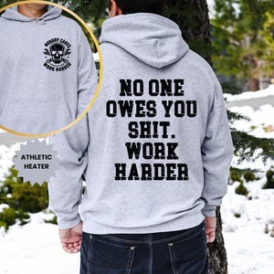 Op de afbeelding: Een lichtgrijze atletische hoodie met de tekst "NO ONE OWES YOU SHIT. WORK HARDER" in het zwart op de achterkant. De voorkant heeft een cirkelvormige afbeelding met een schedel, moersleutels en de woorden "NOBODY CARES WORK HARDER".