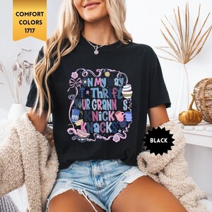 Może przedstawiać: Czarny t-shirt z kolorową grafiką z napisem "On My Way To Thrifting Your Granny's Knick Knacks". Projekt zawiera kokardy, gwiazdki i r&oacute;żne ilustracje. Koszulka jest oznaczona jako "Comfort Colors 1717" i "Black".