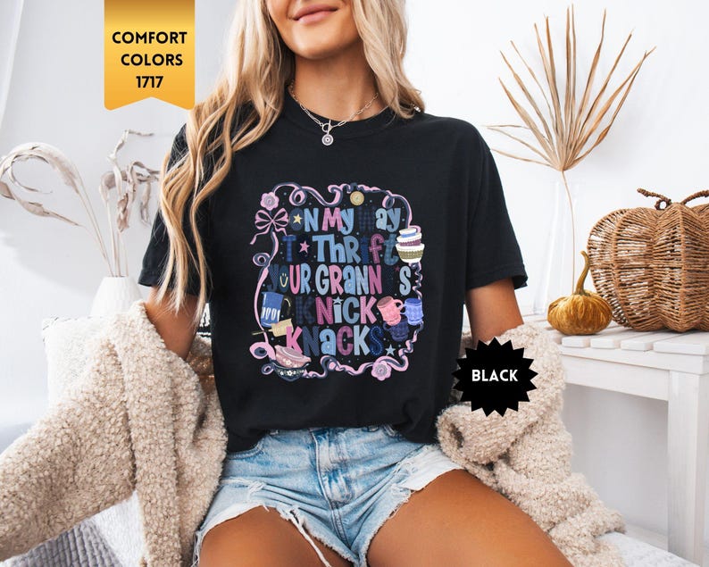 Może przedstawiać: Czarny t-shirt z kolorową grafiką z napisem "On My Way To Thrifting Your Granny's Knick Knacks". Projekt zawiera kokardy, gwiazdki i r&oacute;żne ilustracje. Koszulka jest oznaczona jako "Comfort Colors 1717" i "Black".