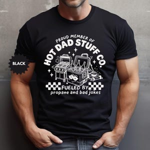 May include: Black t-shirt with a white graphic that reads "Proud Member of Hot Dad Stuff Co." The graphic includes a grill, cooler with bottles, and the text "Fueled by propane and bad jokes."