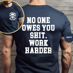 Op de afbeelding: Marineblauw t-shirt met witte tekst op de achterkant: "NO ONE OWES YOU SHIT. WORK HARDER." Een kleinere afbeelding toont de voorkant van het shirt met een schedel- en moersleutelafbeelding en de woorden "NOBODY CARES WORK HARDER."