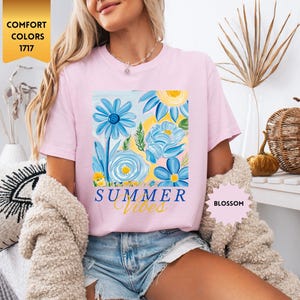 Peut inclure: T-shirt rose avec un motif floral et le texte "Summer Vibes" imprim&eacute; sur le devant. Le motif floral pr&eacute;sente des fleurs bleues, jaunes et roses.