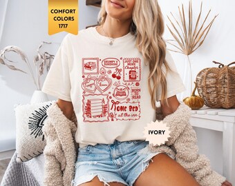 May include: An ivory short-sleeve t-shirt featuring a red graphic print with various book-themed illustrations and text including "Grumpy X Sunshine," "Just One More Chapter," "Enemies to Lovers," "Born to Read," and "One Bed at the Inn."