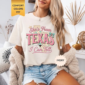May include: An ivory short-sleeve t-shirt featuring a graphic print of the state of Texas with a floral pattern, a small crown, a teal bow, and a longhorn skull. The text reads She's From TEXAS I Can Tell in pink and brown cursive and block lettering.