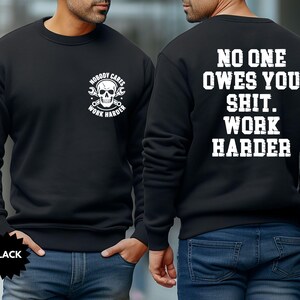 Op de afbeelding: Zwart sweatshirt met witte tekst en afbeeldingen. De voorkant heeft een schedel- en moersleutelontwerp met de woorden "NOBODY CARES WORK HARDER". De achterkant toont "NO ONE OWES YOU SHIT. WORK HARDER" in witte letters.