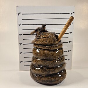 Puede incluir: Un tarro de miel de cerámica con forma de colmena con un cucharón de madera. El tarro es marrón y tiene una superficie texturizada. El cucharón sobresale de la parte superior del tarro.