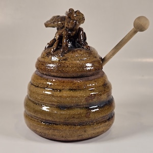 May include: A ceramic honey pot shaped like a beehive with a bee on top and a wooden dipper. The pot is glazed in brown and tan with a glossy finish.