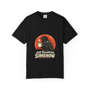 以下が含まれることがあります： コーヒーカップを持ったカラスのグラフィックが特徴の黒いTシャツ。カラスは赤い円を背景に配置され、その下に「Still Breathing Somehow」の文字。普段使いにぴったりのカジュアルなTシャツです。