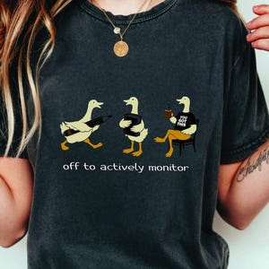 May include: Dark gray t-shirt featuring three cartoon ducks. The ducks are depicted in various poses, one holding a pencil, another with a jacket, and the last one sitting with a coffee cup. The text "off to actively monitor" is printed below the ducks.