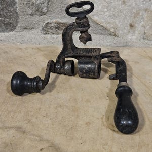 May include: An antique, cast iron tool with a black finish. It features a handle, a crank, and a clamp-like mechanism. The tool is designed for a specific mechanical purpose, likely for shaping or joining materials.