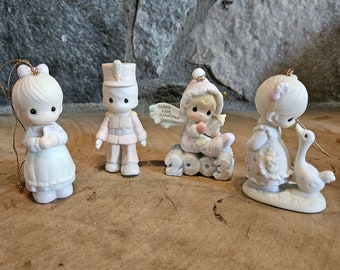 May include: Set of four porcelain Christmas ornaments featuring Precious Moments characters. The ornaments depict a girl holding a letter, a boy in a soldier's uniform, a girl holding a baby, and a girl with a goose.