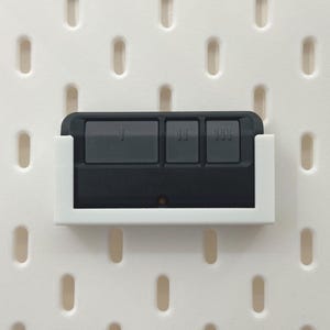 Può includere: Un telecomando nero per l'apertura del garage con tre pulsanti grigi è tenuto in un supporto di plastica bianco. Il supporto è montato su una pannello forato bianco con fori ovali distanziati.