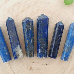 Pode incluir: Sete pontas de cristal de lapis-lazúli azul, cada uma com uma ponta pontiaguda e uma superfície lisa e polida. As pontas estão dispostas em uma fileira sobre uma superfície de madeira marrom claro.