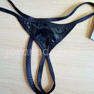 May include: Black thong underwear with a shiny, patterned front panel and thin straps. The edges are trimmed with a darker fabric. The underwear is displayed on a light wood surface.