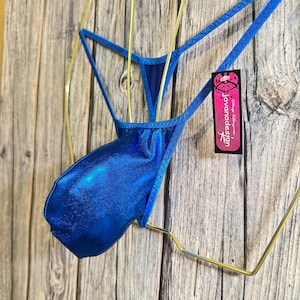 Könnte beinhalten: Ein leuchtend blauer String-Badeanzug mit metallischem Glanz. Das Kleidungsstück hängt an einem goldfarbenen Kleiderbügel vor einem Holzgrund. Ein Etikett mit dem Text "jovanadesign" ist angebracht.