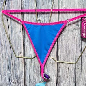 May include: A bright blue thong with vibrant pink trim and thin straps. A silver-toned metal plug with a purple circular gem and a small light blue dolphin charm hangs from the bottom center. A black tag with the text jovanadesign is attached.