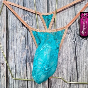 May include: Turquoise lace thong underwear with a peach-colored trim, displayed on a gold-colored hanger. The underwear features a delicate lace pattern. The tag reads "jovanadesign".