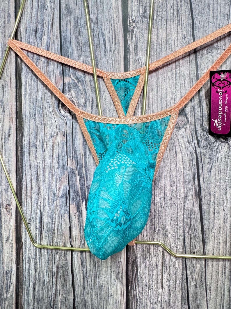May include: Turquoise lace thong underwear with a peach-colored trim, displayed on a gold-colored hanger. The underwear features a delicate lace pattern. The tag reads "jovanadesign".