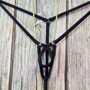 May include: Black thong underwear with a unique design. The thong features a series of black straps connected by small metal rings. The design includes a loop in the front. The background is a wooden surface.