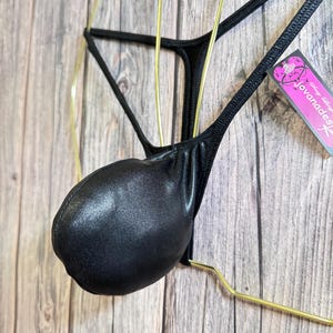 May include: Black thong-style underwear with a pouch, displayed on a gold-colored hanger. The underwear has a shiny, textured appearance. A tag with the text "Jovanadesign" is visible.