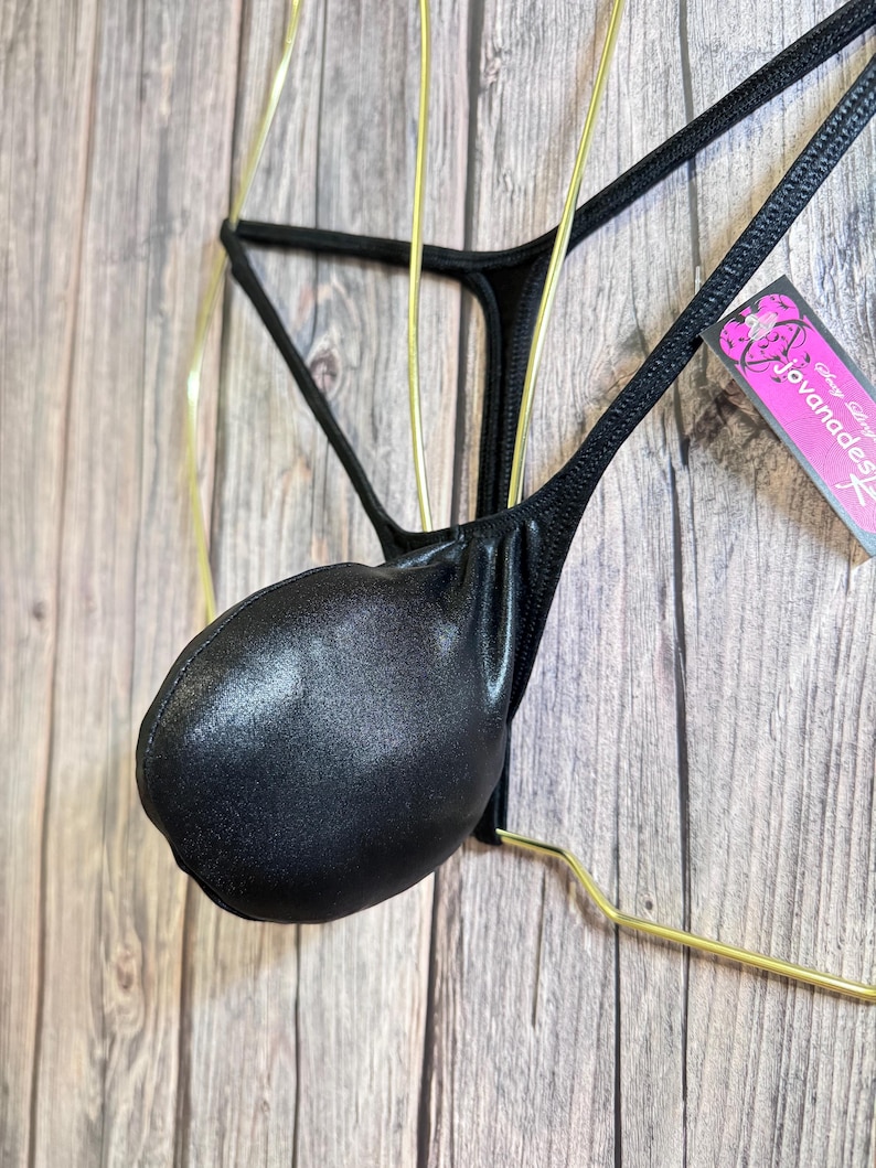 May include: Black thong-style underwear with a pouch, displayed on a gold-colored hanger. The underwear has a shiny, textured appearance. A tag with the text "Jovanadesign" is visible.