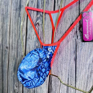 May include: A thong-style undergarment with a vibrant blue and white patterned pouch, outlined with bright red trim. The garment is displayed on a gold-colored hanger, with a tag that reads "jovanadesign". The background is a weathered wooden surface.