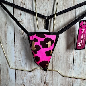 May include: A thong featuring a vibrant neon pink fabric with a black and gold leopard print pattern. The garment has thin black elastic straps and a black trim. A rectangular tag attached to the side displays the text jovanadesign.