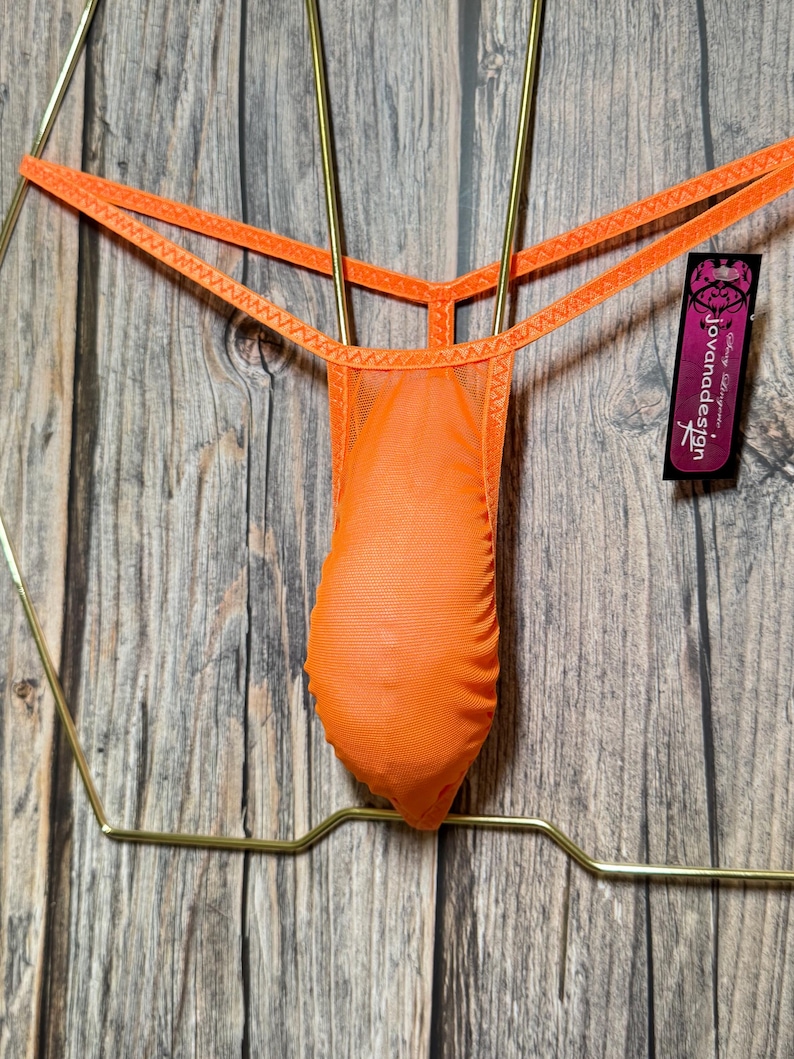 Op de afbeelding: Oranje mesh string ondergoed hangend aan een gouden hanger. Het ondergoed heeft een jovanadesign-label.