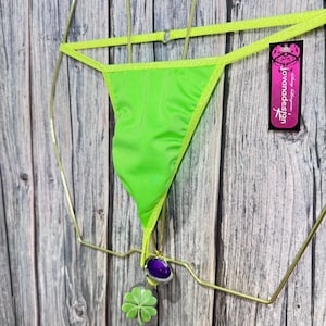 May include: Neon green thong bikini with a matching trim. A purple gem and a green four-leaf clover charm dangle from the bottom. The bikini is displayed on a gold-colored hanger against a wooden background. A tag with the text "jovanadesign" is visible.