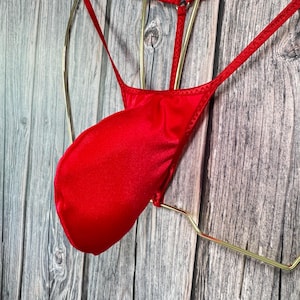 Könnte beinhalten: Ein leuchtend roter String-Tanga aus glattem, glänzendem Stoff. Das Unterteil hat eine dreieckige Vorderseite und schmale Träger. Es wird auf einem goldfarbenen Kleiderbügel vor einem Holzgrund präsentiert.