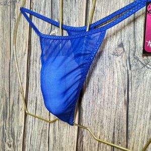 May include: A sheer blue thong with delicate straps. The lingerie is displayed on a gold-colored hanger against a wooden background. The fabric appears lightweight and slightly transparent.