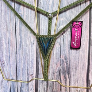 May include: An olive green thong featuring thin elastic straps with a zigzag stitch pattern and a central triangular panel with a metallic blue and green iridescent finish. A black tag with the text jovanadesign is attached to the side.