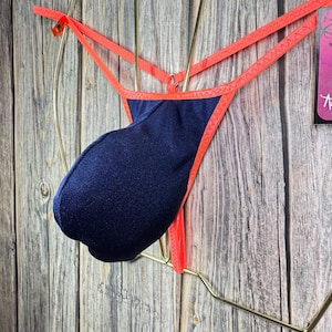 Könnte beinhalten: Ein marineblauer Tanga mit einem leuchtend orangenen Saum. Der Tanga hängt an einem goldenen Kleiderbügel vor einem Holz Hintergrund.