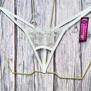 May include: A light green thong with a floral lace panel. The lingerie features a delicate design with thin straps and is displayed on a gold-colored hanger. A tag with the text "jovanadesign" is attached.
