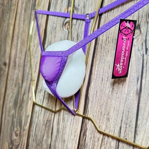 Könnte beinhalten: Ein violetter String-Tanga mit Mesh-Front und dünnen Trägern. Das Kleidungsstück wird auf einem weißen, kugelförmigen Objekt präsentiert, das in einem goldfarbenen Drahtrahmen aufgehängt ist. Ein Etikett mit dem Text "jovanadesign" ist angebracht.