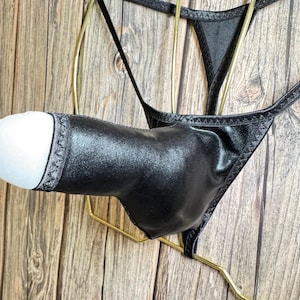 Puede incluir: Tanga negro con una bolsa, exhibido en una percha dorada. La ropa interior está hecha de un material oscuro y brillante con un ribete decorativo. El fondo es una superficie de madera.