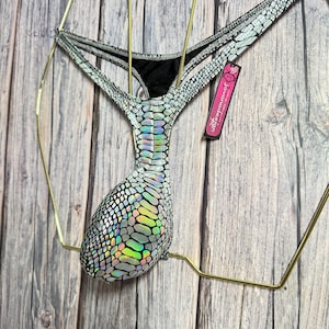 Könnte beinhalten: Silberner und schwarzer String-Bikini mit Schlangenmuster. Der Bikini wird auf einem goldenen Drahtbügel vor einem Holzgrund gezeigt. Der Bikini hat ein schwarzes Futter und ein Etikett mit der Aufschrift "jovandesign".