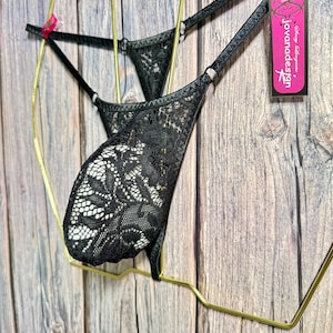 Puede incluir: Tanga de encaje negro con tirantes ajustables. La lencer&iacute;a presenta un patr&oacute;n floral de encaje y se exhibe en una percha dorada. Una etiqueta rosa con el texto "jovanadesign" es visible.