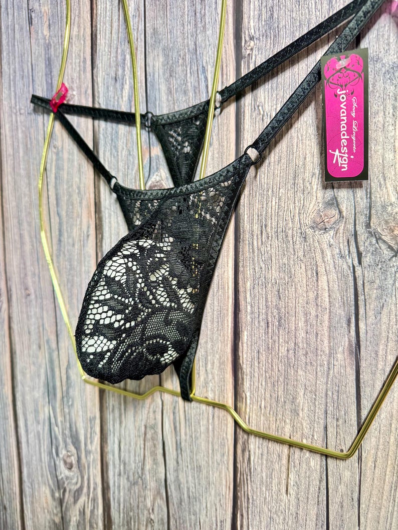 Puede incluir: Tanga de encaje negro con tirantes ajustables. La lencer&iacute;a presenta un patr&oacute;n floral de encaje y se exhibe en una percha dorada. Una etiqueta rosa con el texto "jovanadesign" es visible.