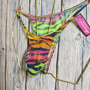 May include: A vibrant, multi-colored thong bikini bottom with a zebra stripe pattern in neon green, pink, and orange. The garment is displayed on a gold-colored hanger against a wooden background. A tag with the text "jovanadesign" is attached.