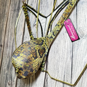 May include: A thong-style swimsuit with a snakeskin pattern in shades of yellow, green, and black. The garment is displayed on a gold-colored hanger against a wooden background. A pink tag with the text "Jovanadesign" is attached.